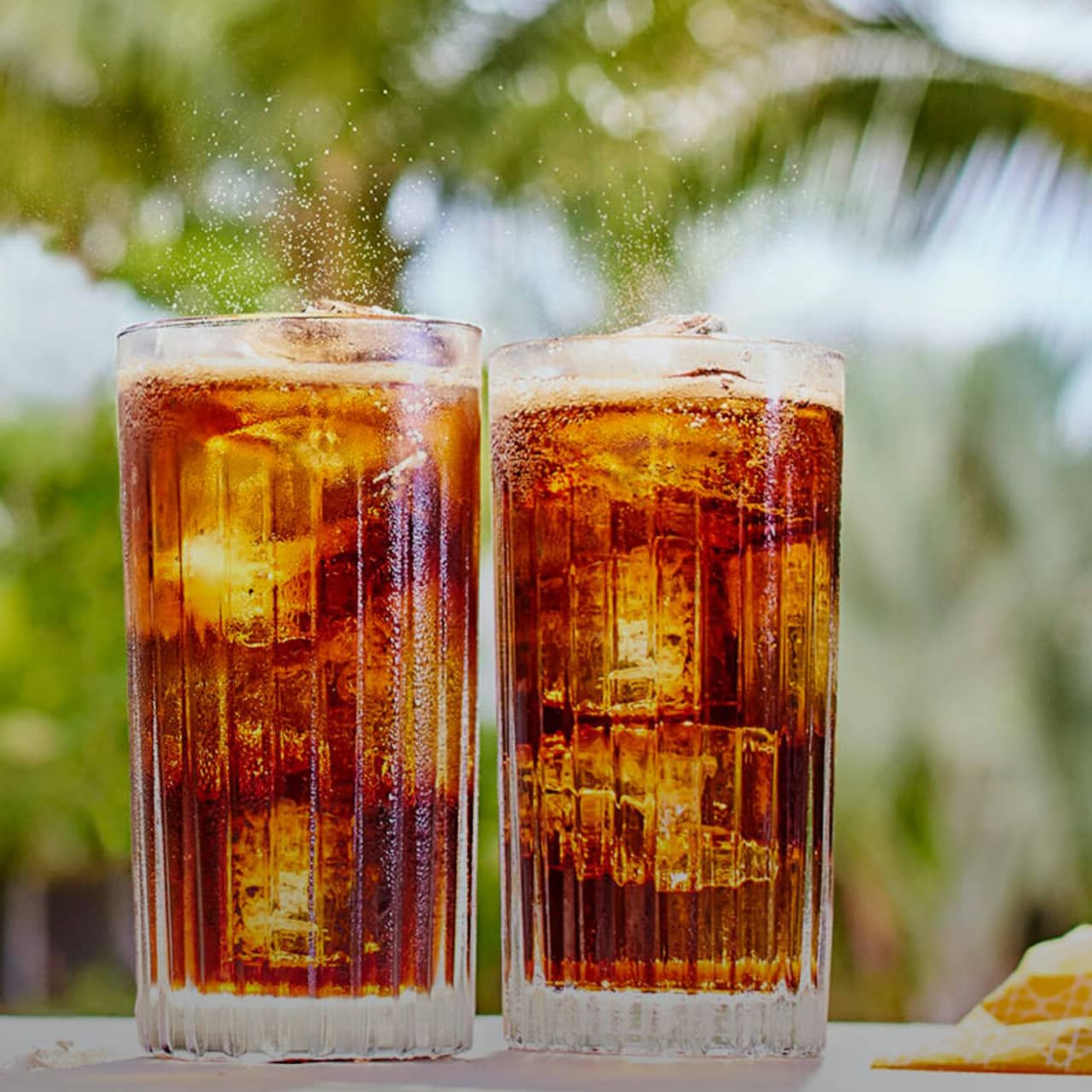 Two tall glasses with dark fizzy drinks, ice cubes, and a lemon wedge.