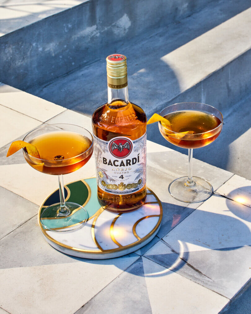 Bacardí Añejo Cuatro rum bottle with two cocktail-filled coupe glasses.