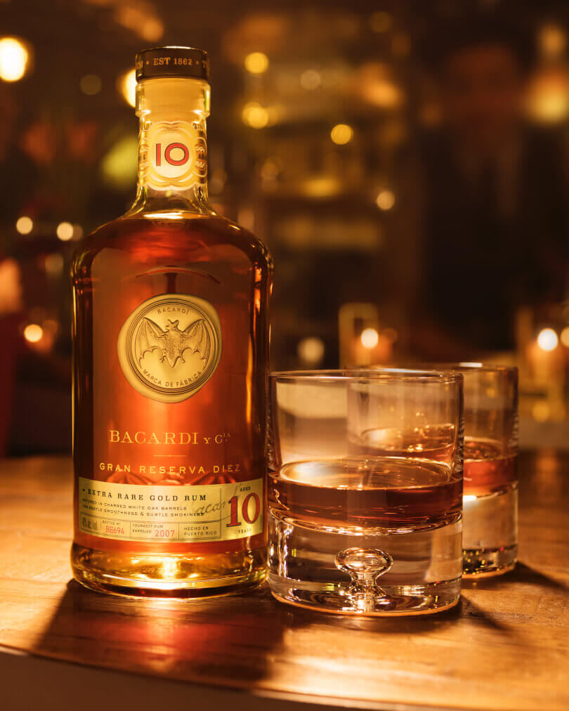 BACARDÍ Gran Reserva Diez rum bottle next to two glasses with amber liquid.
