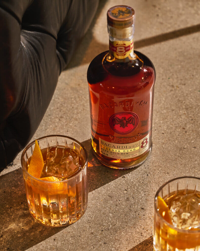 BACARDÍ Reserva Ocho rum bottle with two cocktail glasses containing amber liquid.