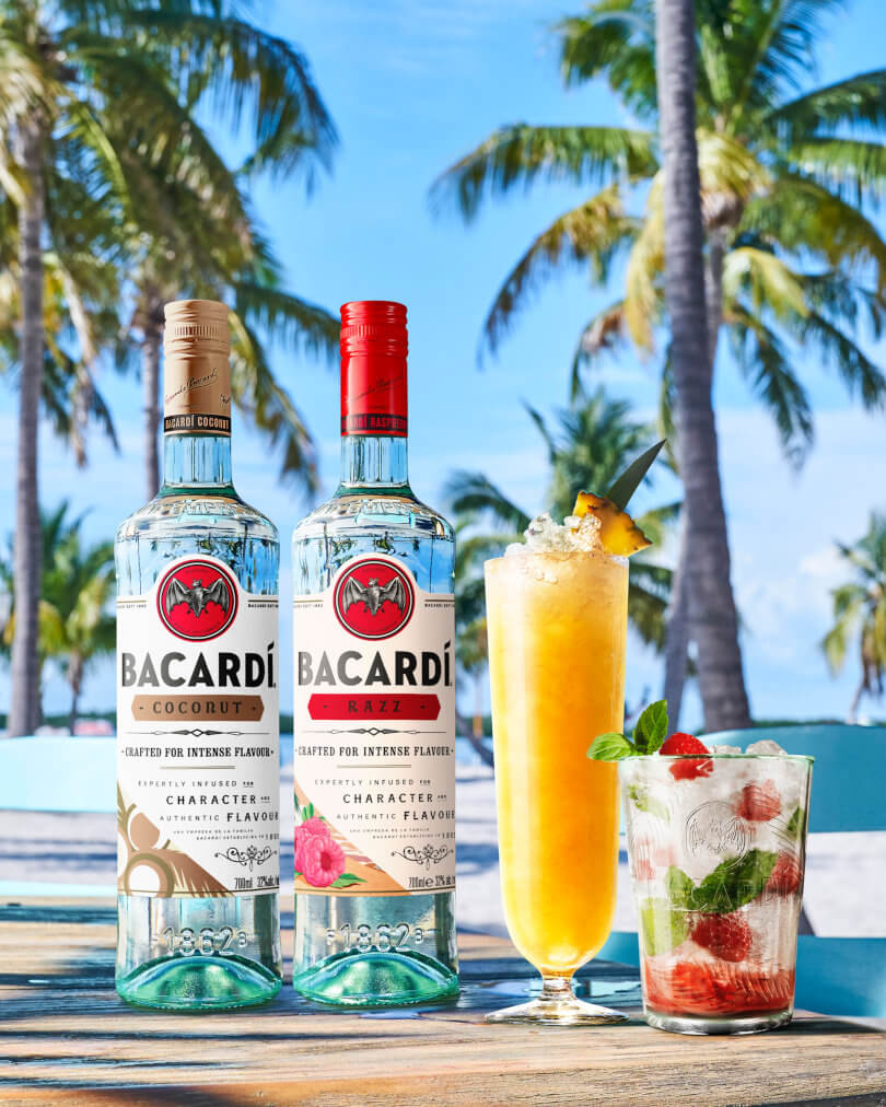 Two BACARDÍ rum bottles, coconut and raspberry flavors, with two cocktails nearby.