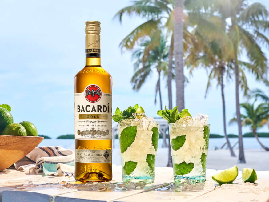 BACARDÍ Gold rum bottle beside two cocktails garnished with mint, lime, and ice cubes.