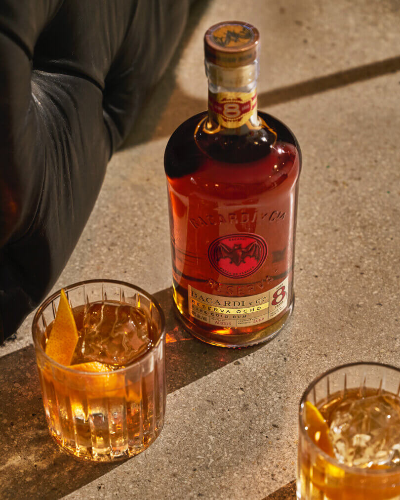 BACARDÍ Reserva Ocho rum bottle with two rocks glasses filled with amber cocktails and orange peels.