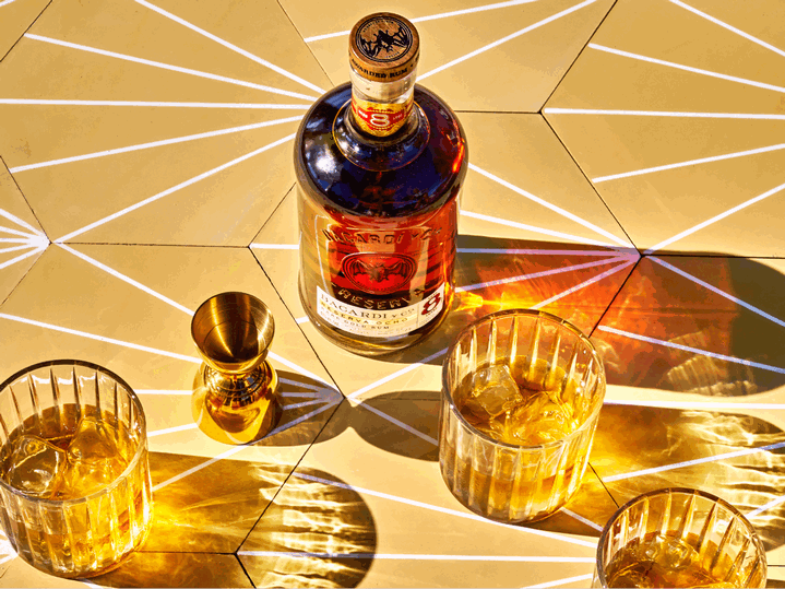 BACARDÍ Reserva Ocho rum bottle with three glasses of golden liquid, ice, and a golden jigger.