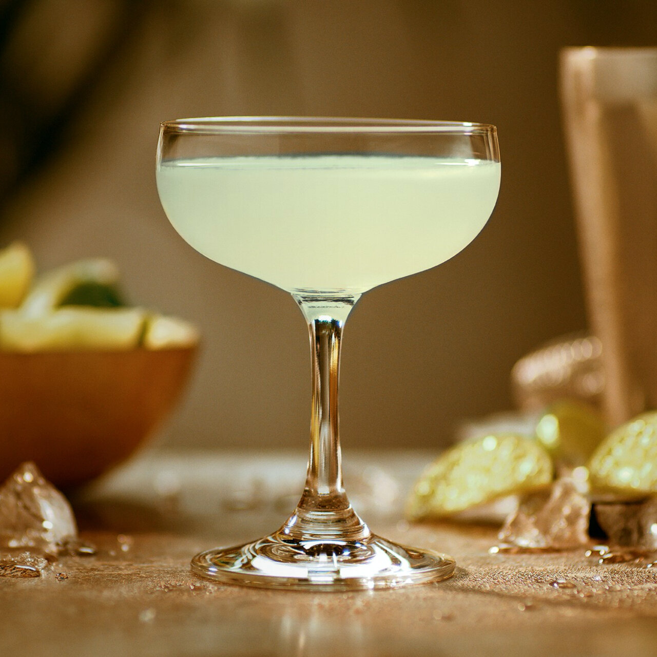 Pale yellow Daiquiri cocktail in a coupe glass with lime wedges and ice cubes.