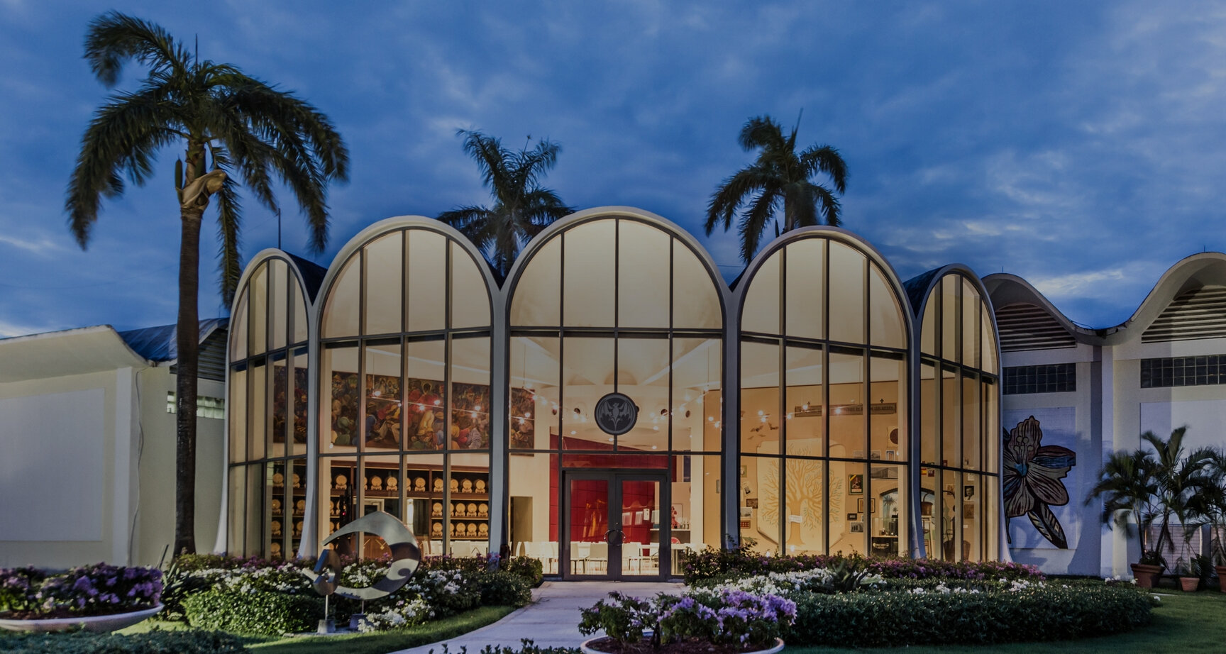 Modern building with arched glass facade, illuminated at dusk, surrounded by lush green gardens and palm trees.