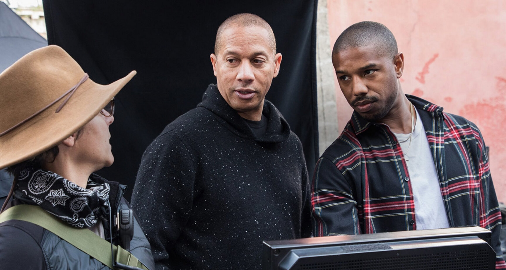 Three people on a set, one wearing a wide-brimmed hat, another in a hoodie, third in plaid shirt.