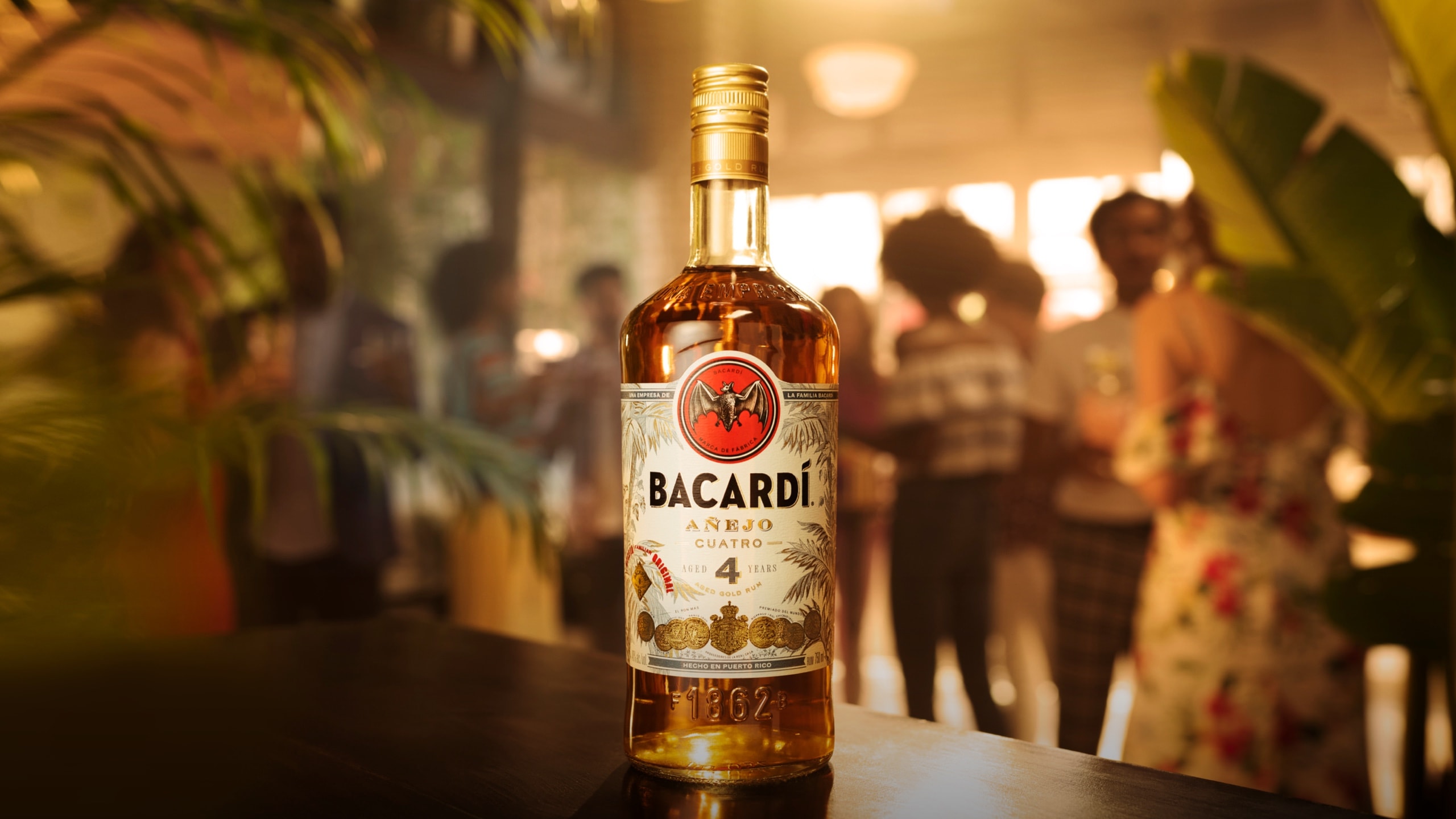 BACARDÍ Añejo Cuatro rum bottle positioned with tropical plants and social gathering in background.