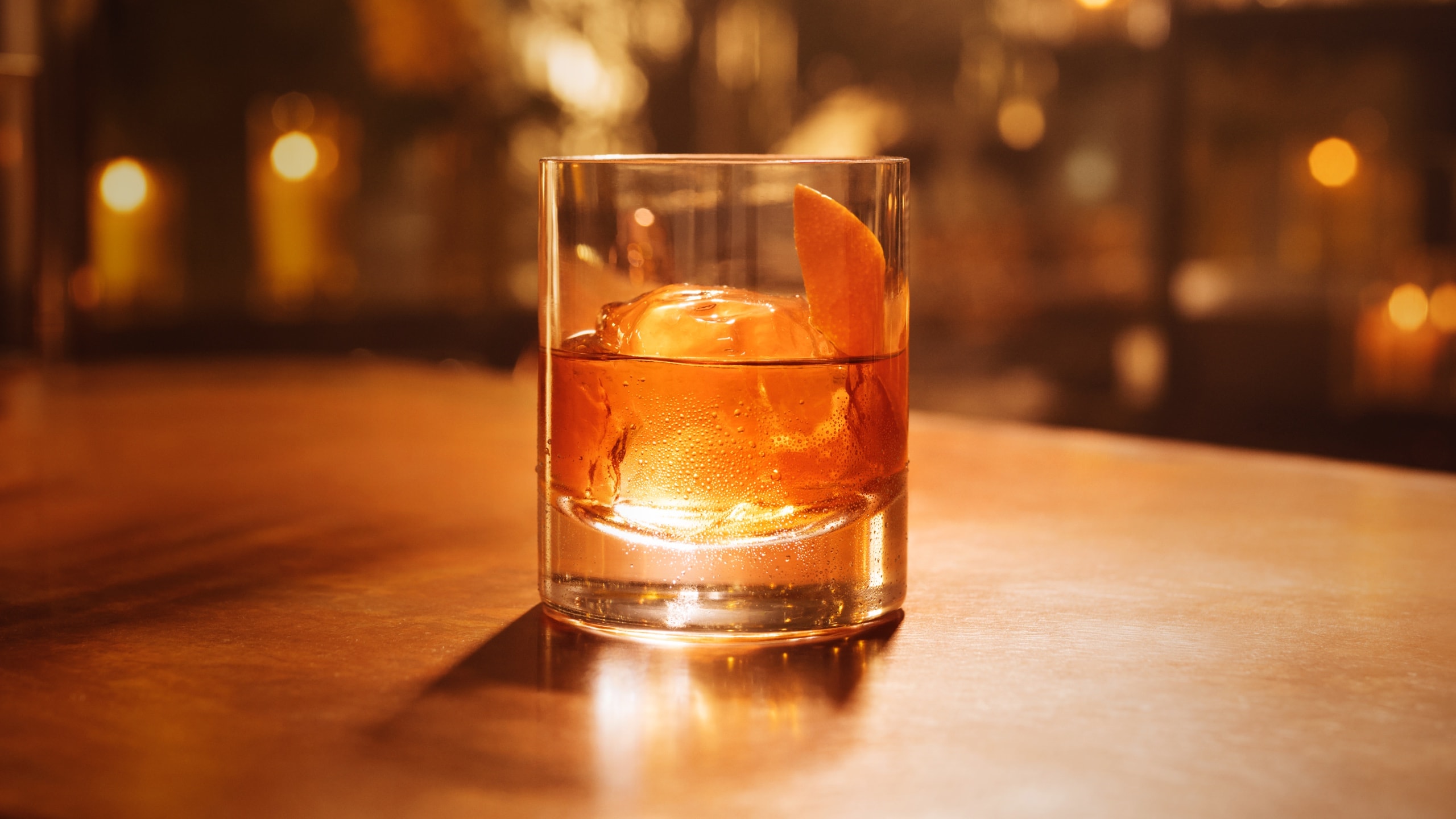 Glass of amber cocktail with large spherical ice cube and orange peel garnish.