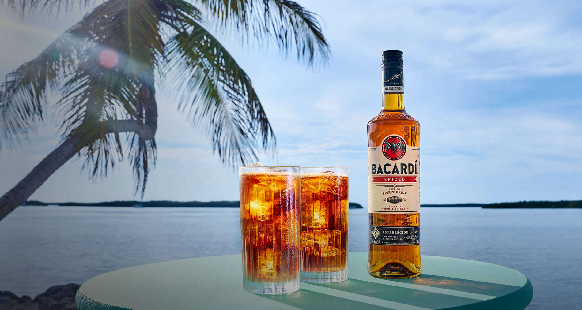Bacardí Spiced rum bottle next to two tall glasses filled with dark iced cocktails.