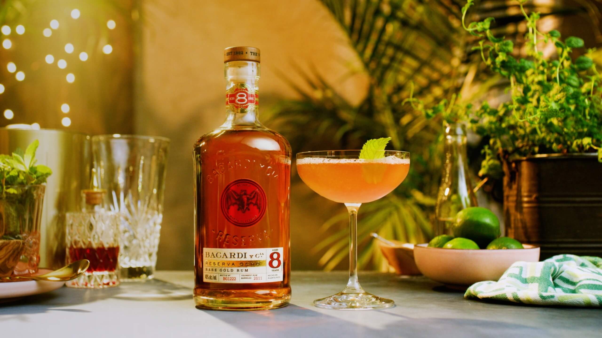 BACARDÍ Reserva Ocho rum bottle next to a coupe glass with amber cocktail, garnished with fresh mint leaf.