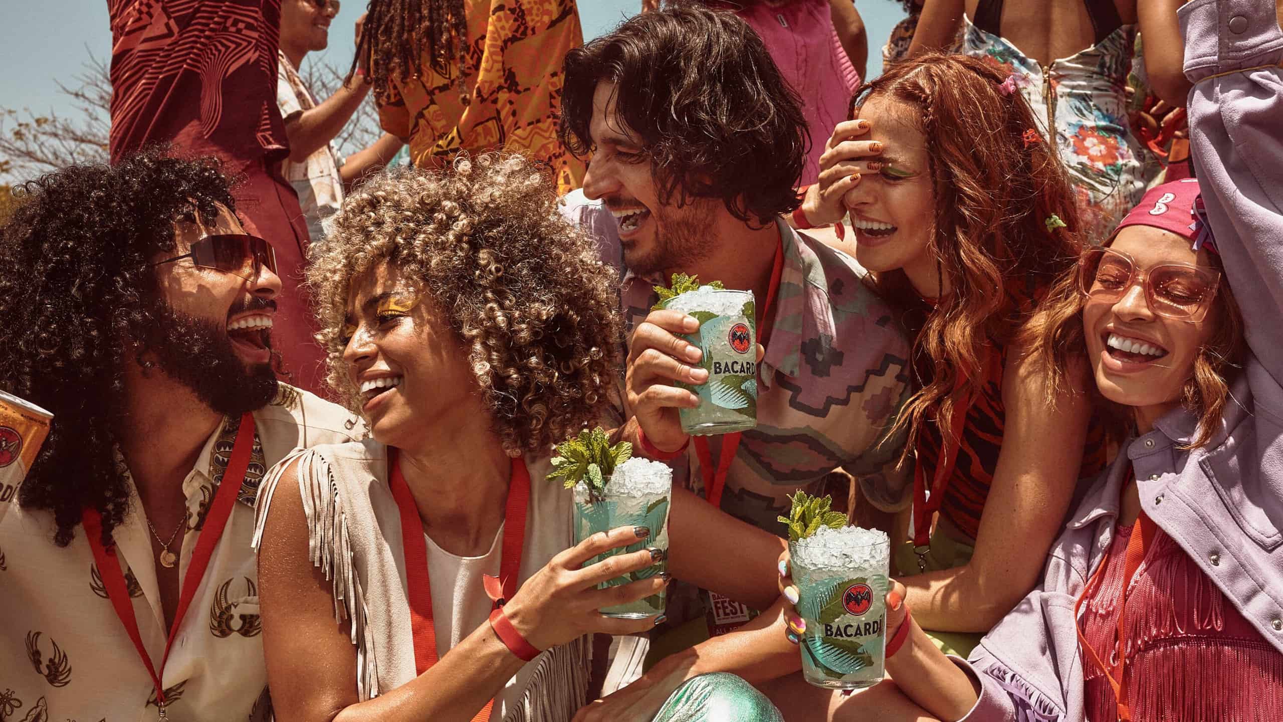 Five people holding BACARDÍ mojito cocktails, laughing and socializing in an outdoor setting.