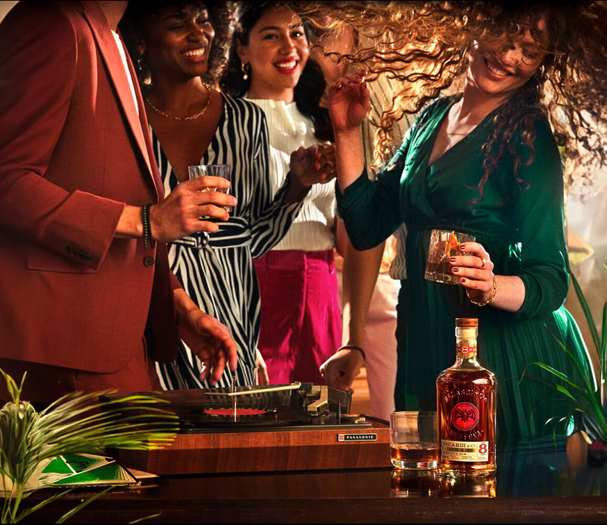 BACARDÍ Reserva 8 rum bottle near turntable with people socializing in background.