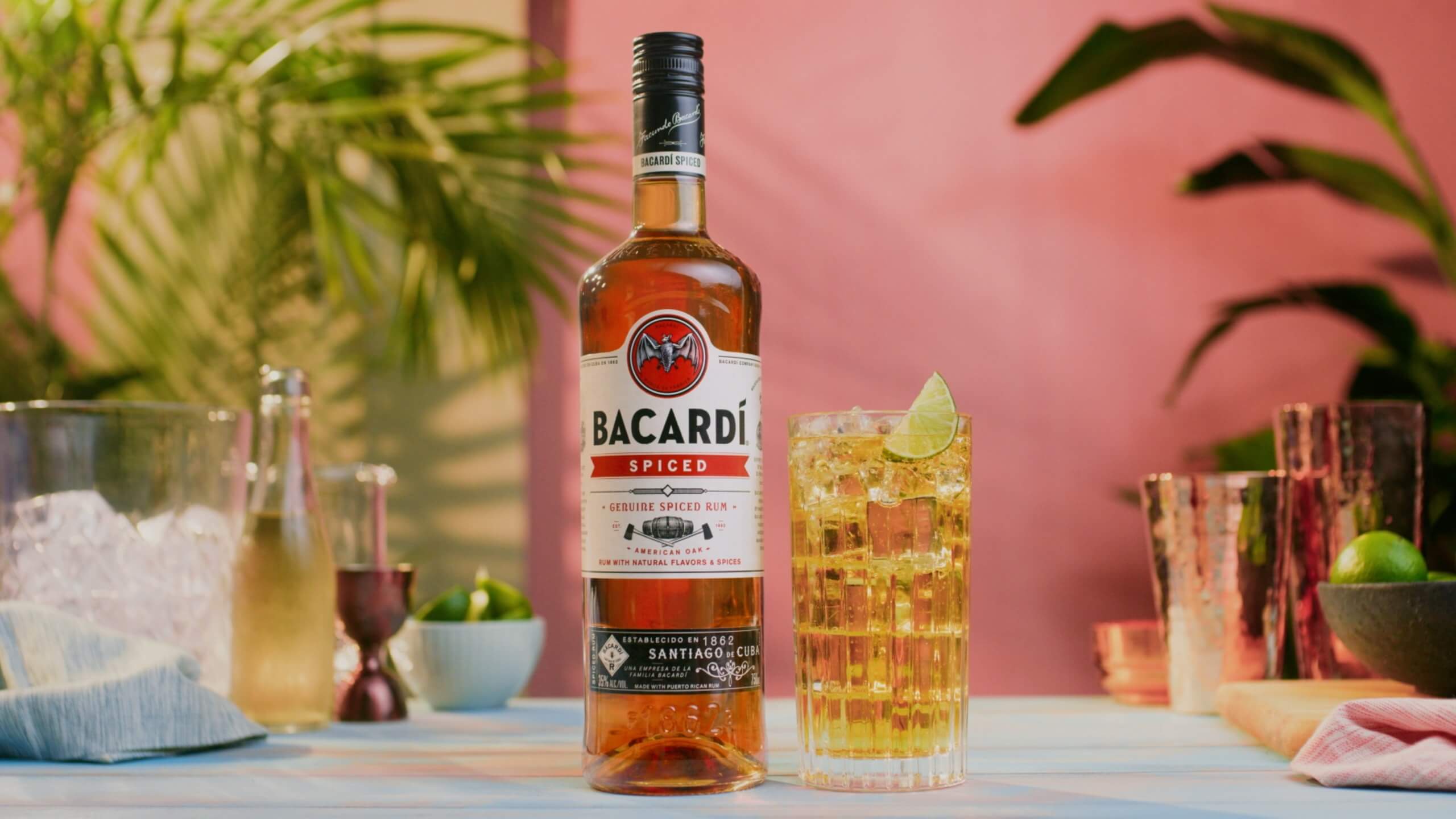 BACARDÍ Spiced rum bottle next to a tall cocktail glass with lime wedge, ice cubes, bar tools nearby.