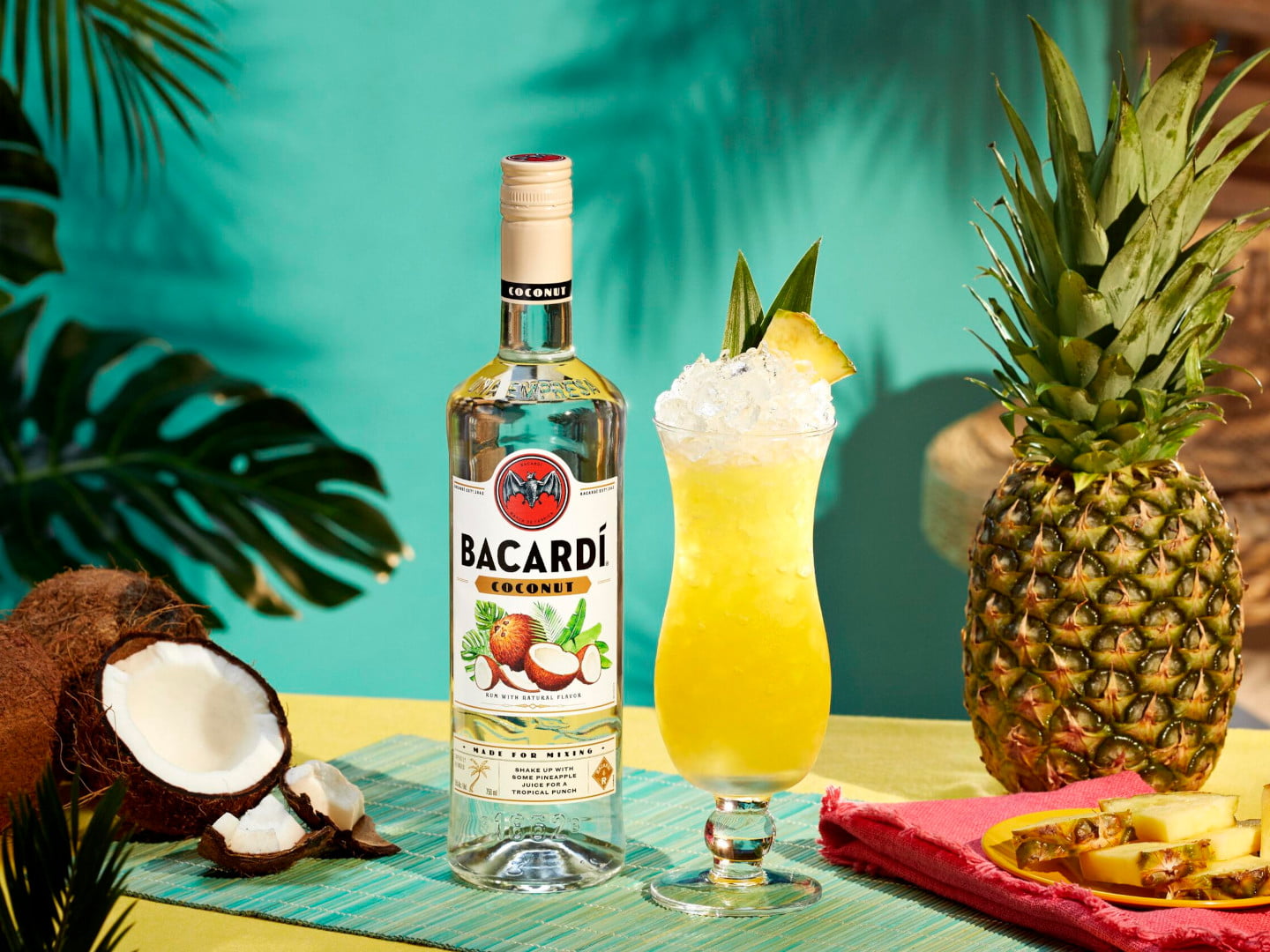 Coconut_Lifestyle BACARDÍ Coconut rum bottle beside a yellow cocktail garnished with pineapple, mint, and a halved coconut.
