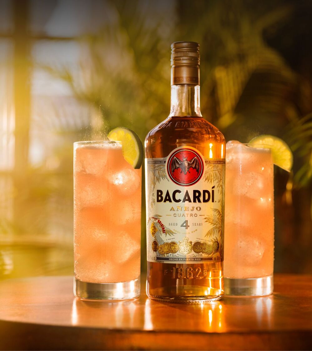 BACARDÍ Añejo Cuatro rum bottle with two tall cocktail glasses, lime wedges, ice.