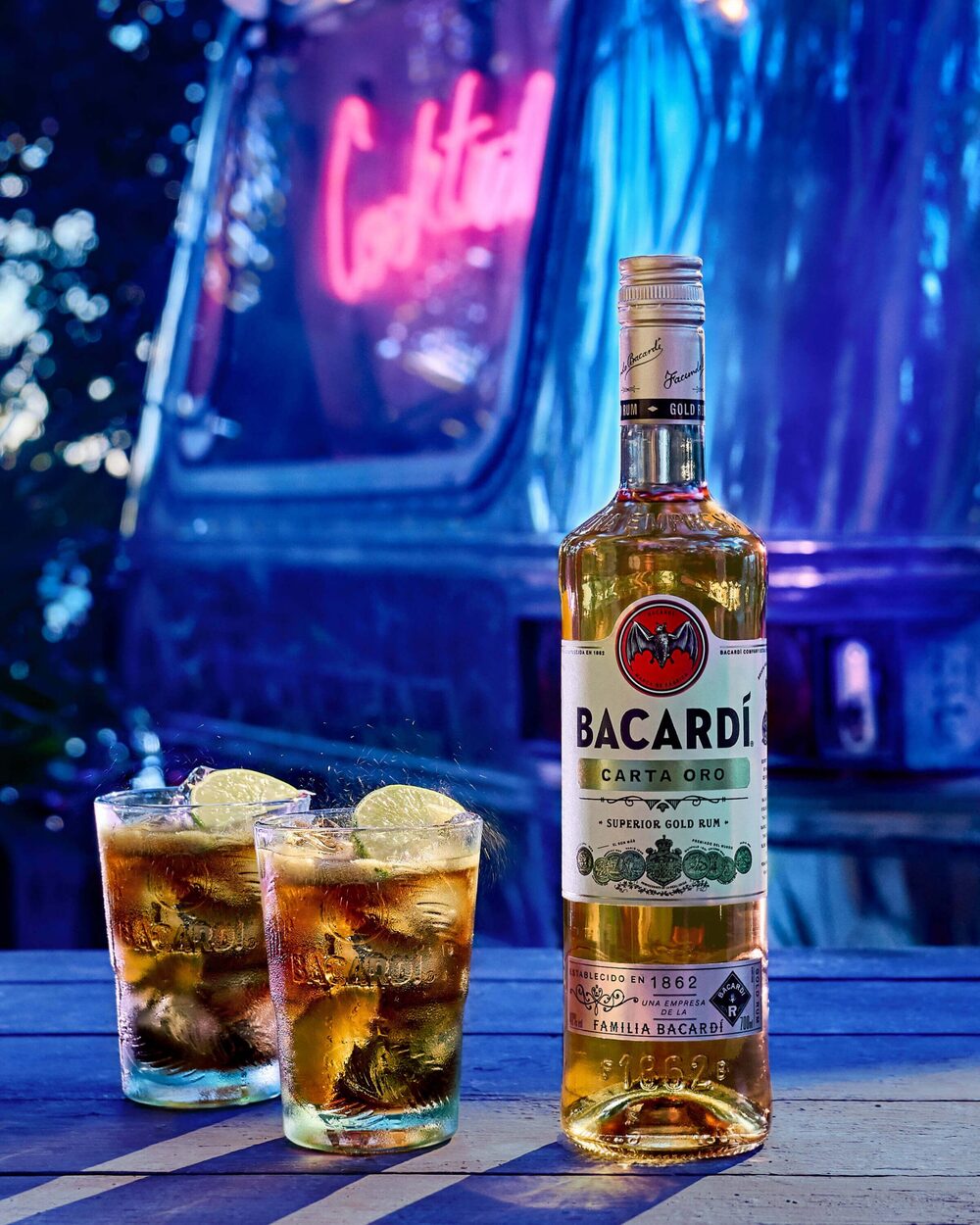BACARDÍ Carta Oro rum bottle next to two cocktail glasses with splashing liquid, lime wedges.