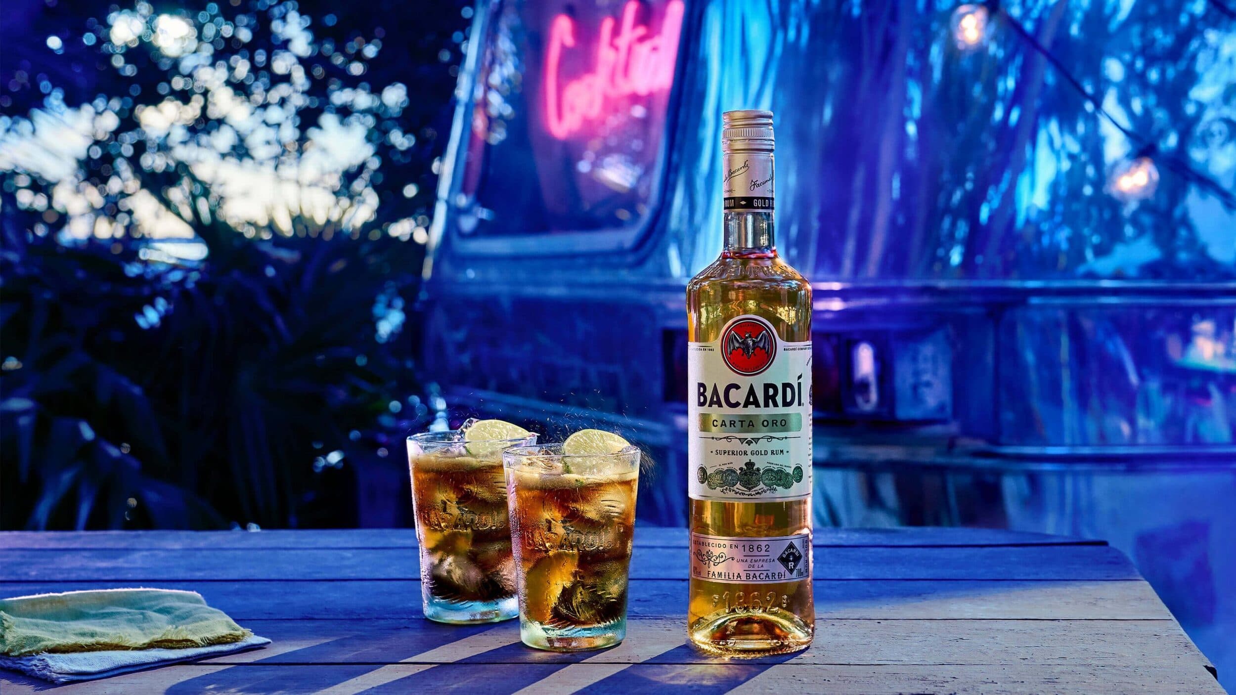 Bacardí Carta Oro rum bottle beside two cocktails with lime wedges and ice.