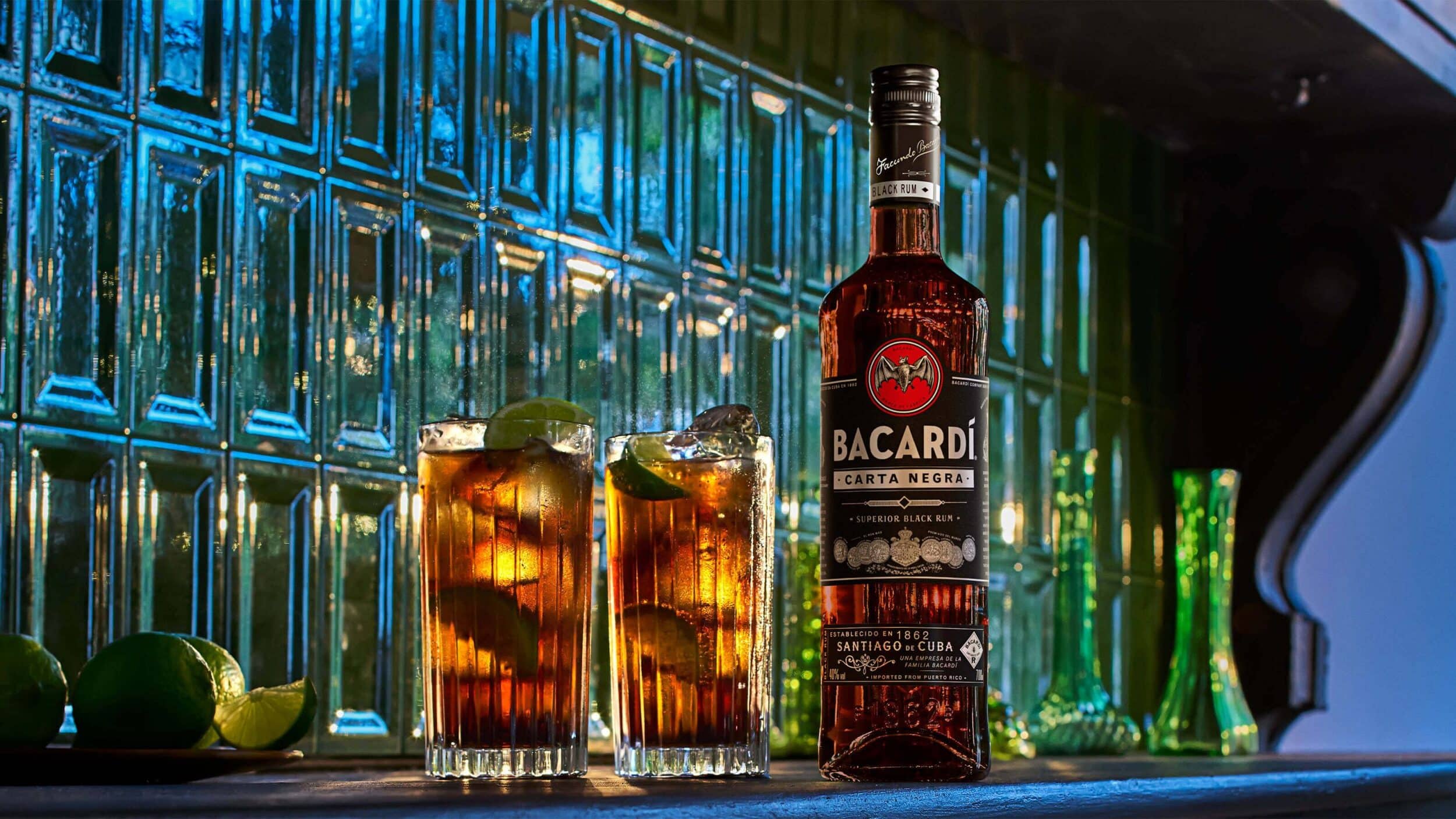 BACARDÍ Carta Negra rum bottle beside two tall glasses with dark cocktails, lime wedges, ice cubes.