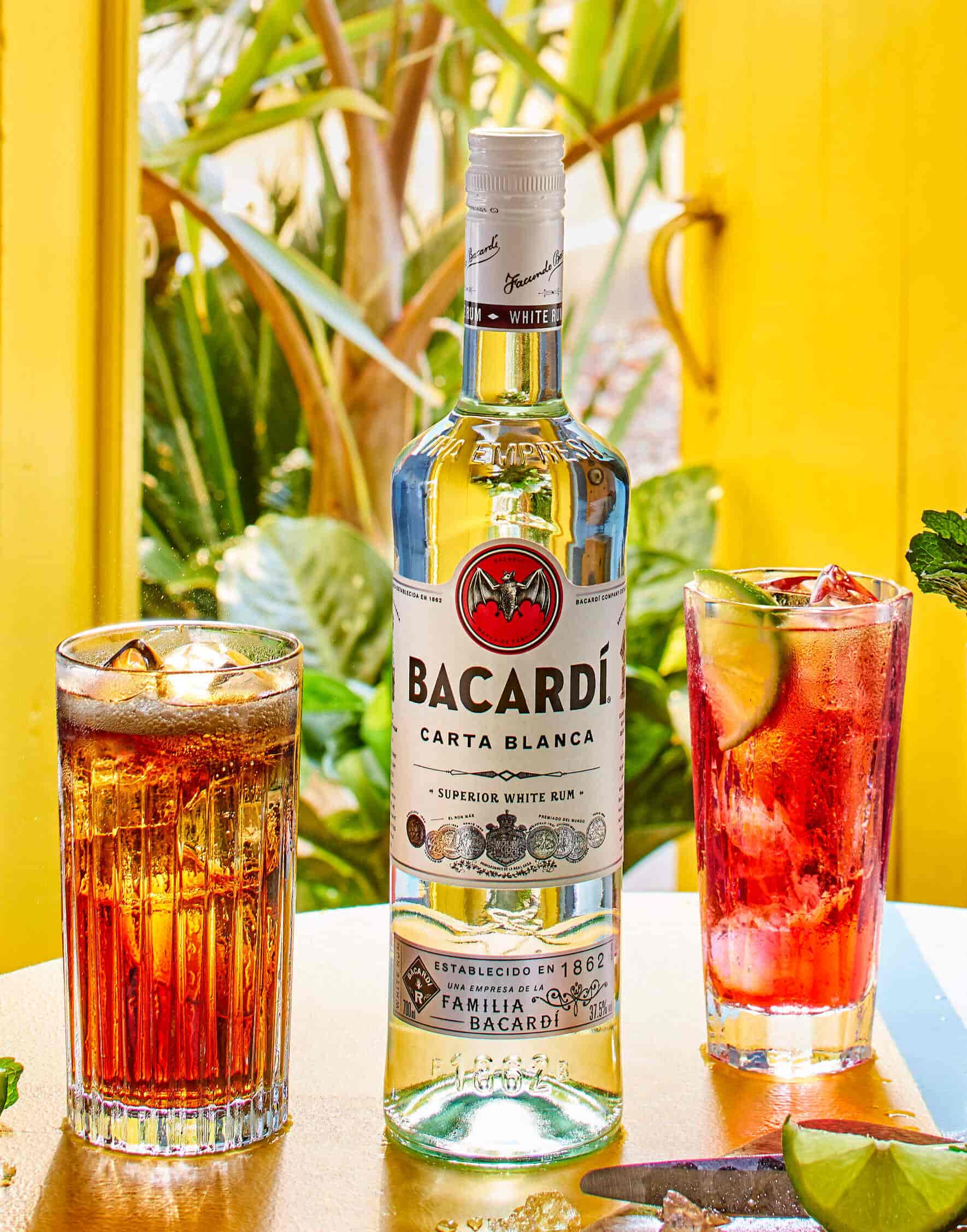 BACARDÍ Carta Blanca rum bottle positioned between two cocktail glasses with dark and red drinks.