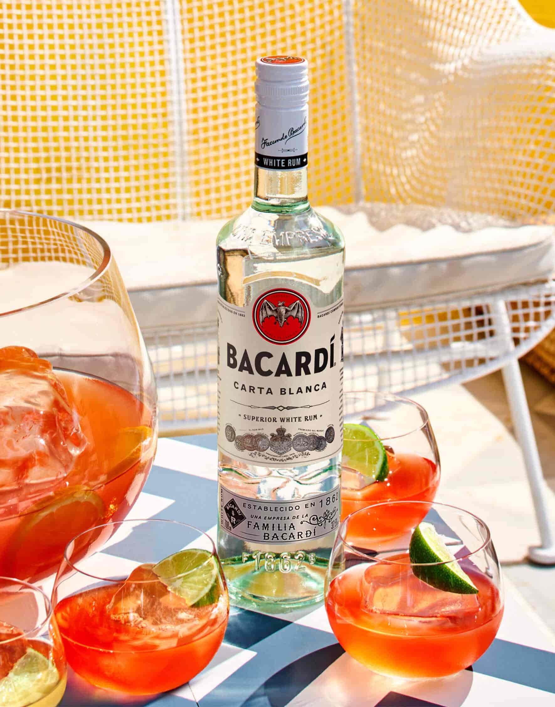FAQ-03-Cola_uk BACARDÍ Carta Blanca rum bottle with cocktail glasses, punch bowl, and lime wedges.