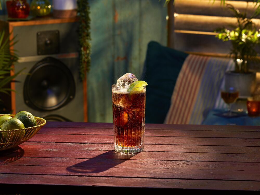 Tall glass of Cuba Libre cocktail with ice cubes and lime wedge, filled with dark, bubbly beverage.