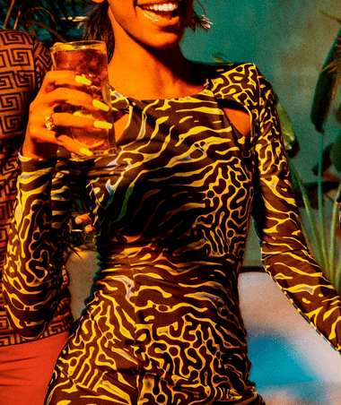 Person holding a tall cocktail glass with a lime wedge, wearing a bold yellow and black patterned dress.