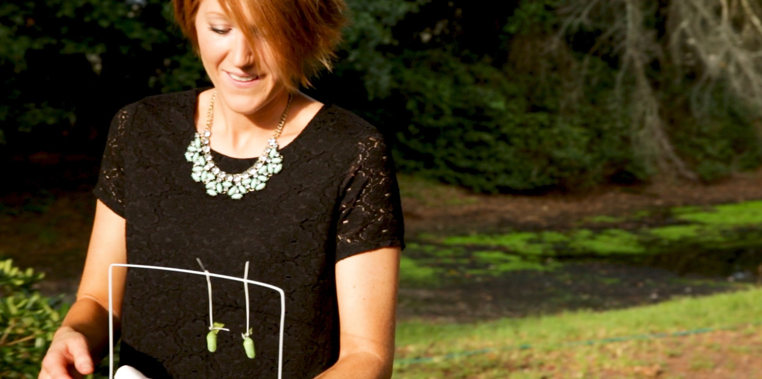 Person holding a white frame with two green objects in an outdoor garden setting.