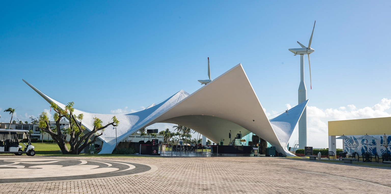 Modern white tensile structure with curved roof, situated on paved ground with a wind turbine and tropical trees.