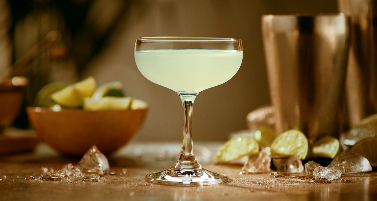 Light green Daiquiri cocktail in a coupe glass with scattered lime wedges and ice.