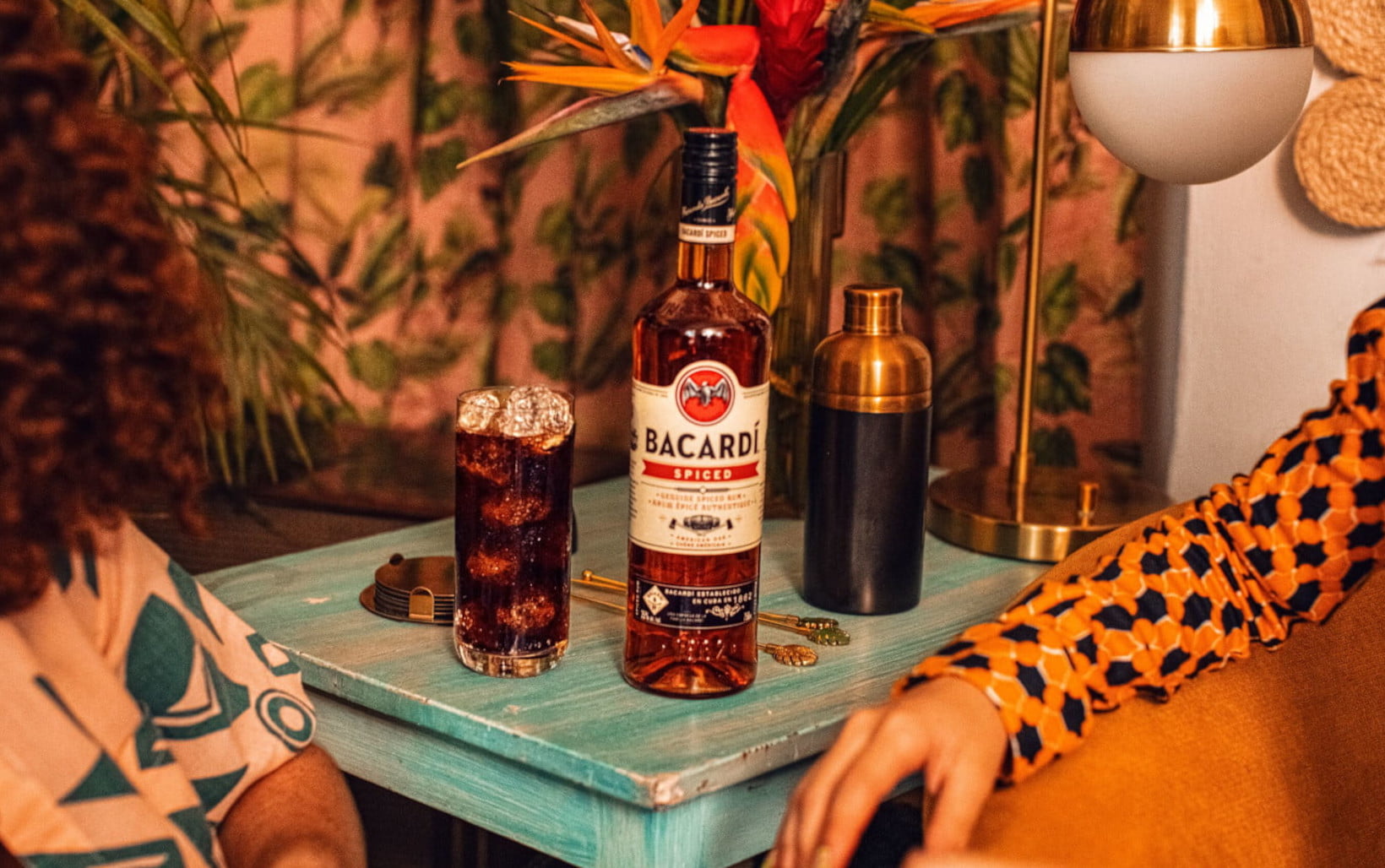 BACARDÍ Spiced rum bottle beside a cola glass, cocktail shaker, with tropical flowers in view.