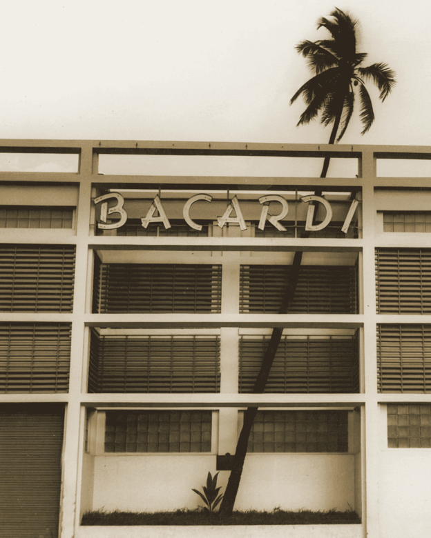 Sepia-toned photograph of BACARDÍ building facade with a leaning palm tree behind the structure.