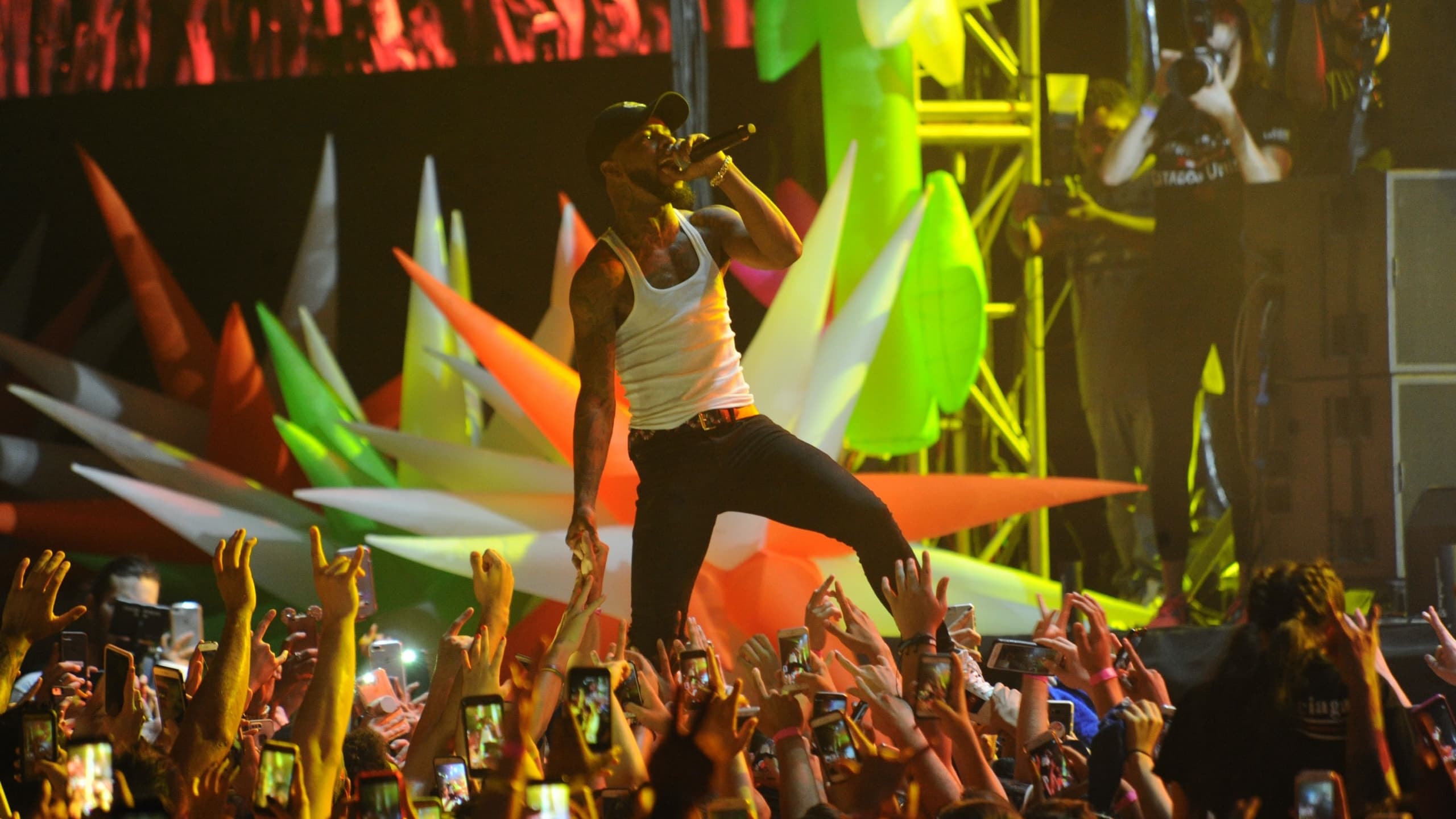 Performer singing on stage with colorful inflatable props, crowd holding up cell phones below.