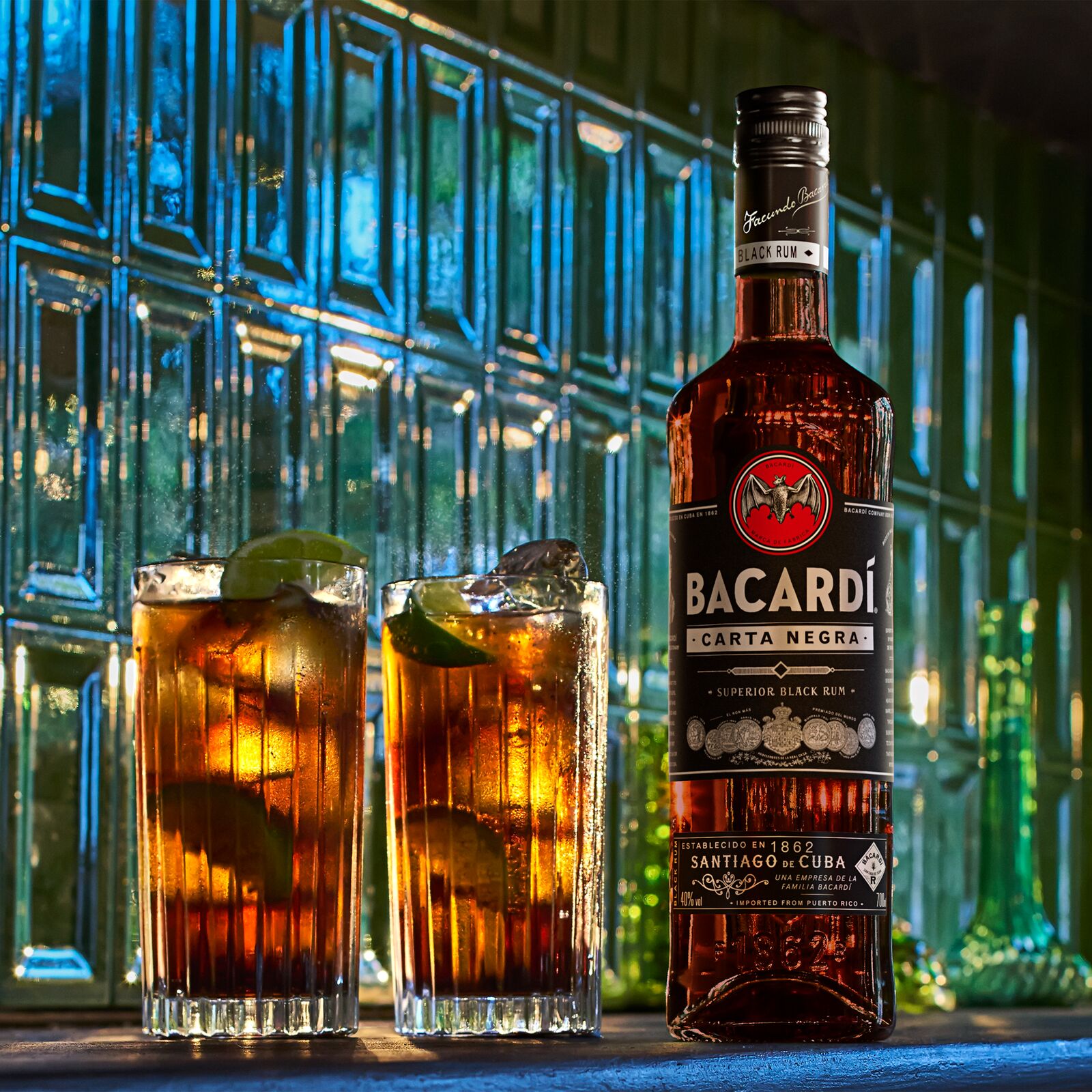 Bacardí Carta Negra rum bottle next to two glasses with dark cocktails, lime wedges, ice.