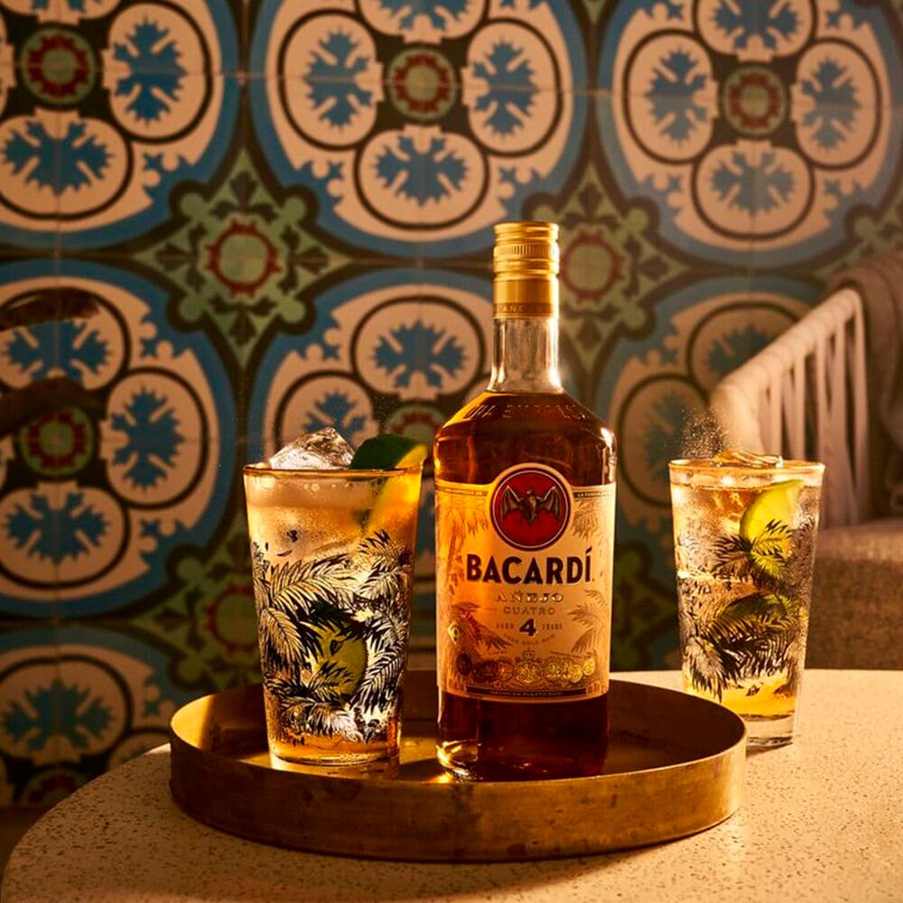 BACARDÍ Añejo Cuatro rum bottle with two cocktails, lime wedges, ice on golden tray.