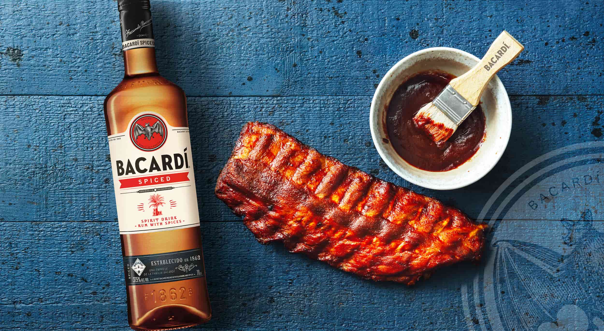 BACARDÍ Spiced Rum bottle positioned near a rack of caramelized BBQ ribs with a dark sauce bowl.