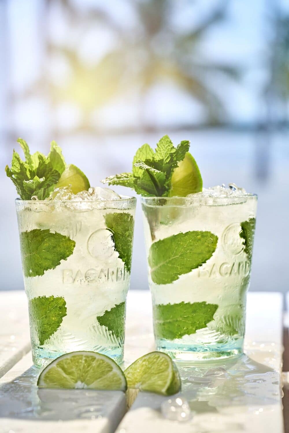 Two Mojito cocktails in embossed glasses with crushed ice, mint leaves, lime wedges.