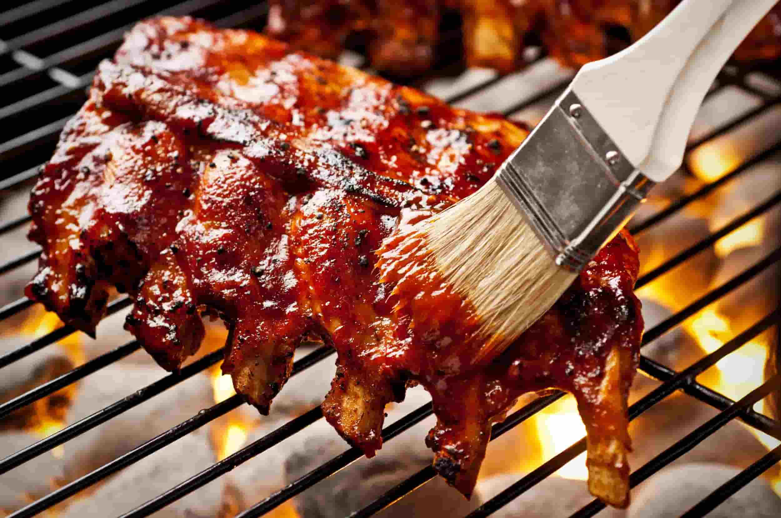 Rack of ribs being basted with sauce while cooking on a grill with glowing embers and flames.
