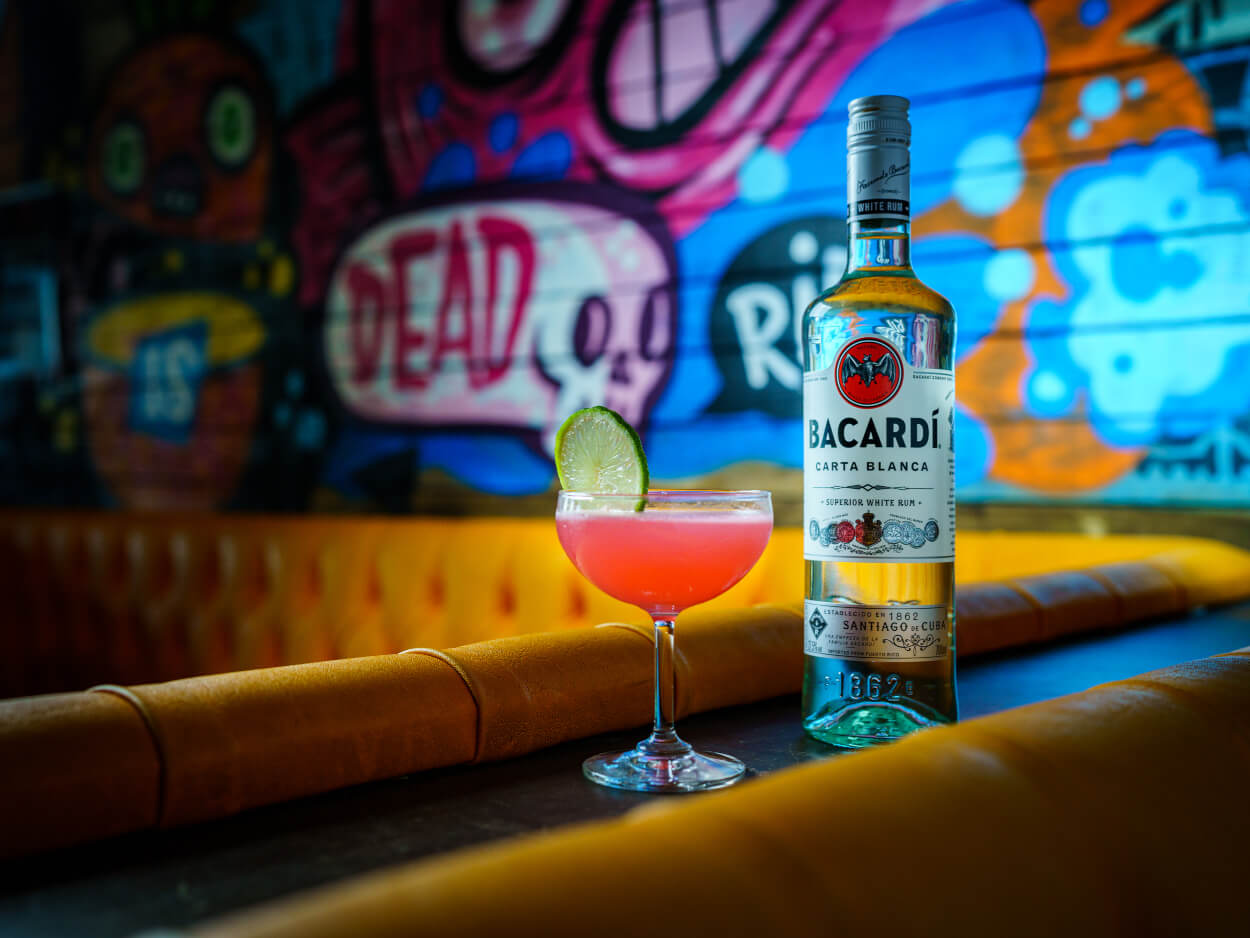 BACARDÍ Carta Blanca rum bottle beside a pink cocktail with lime wedge in a coupe glass.