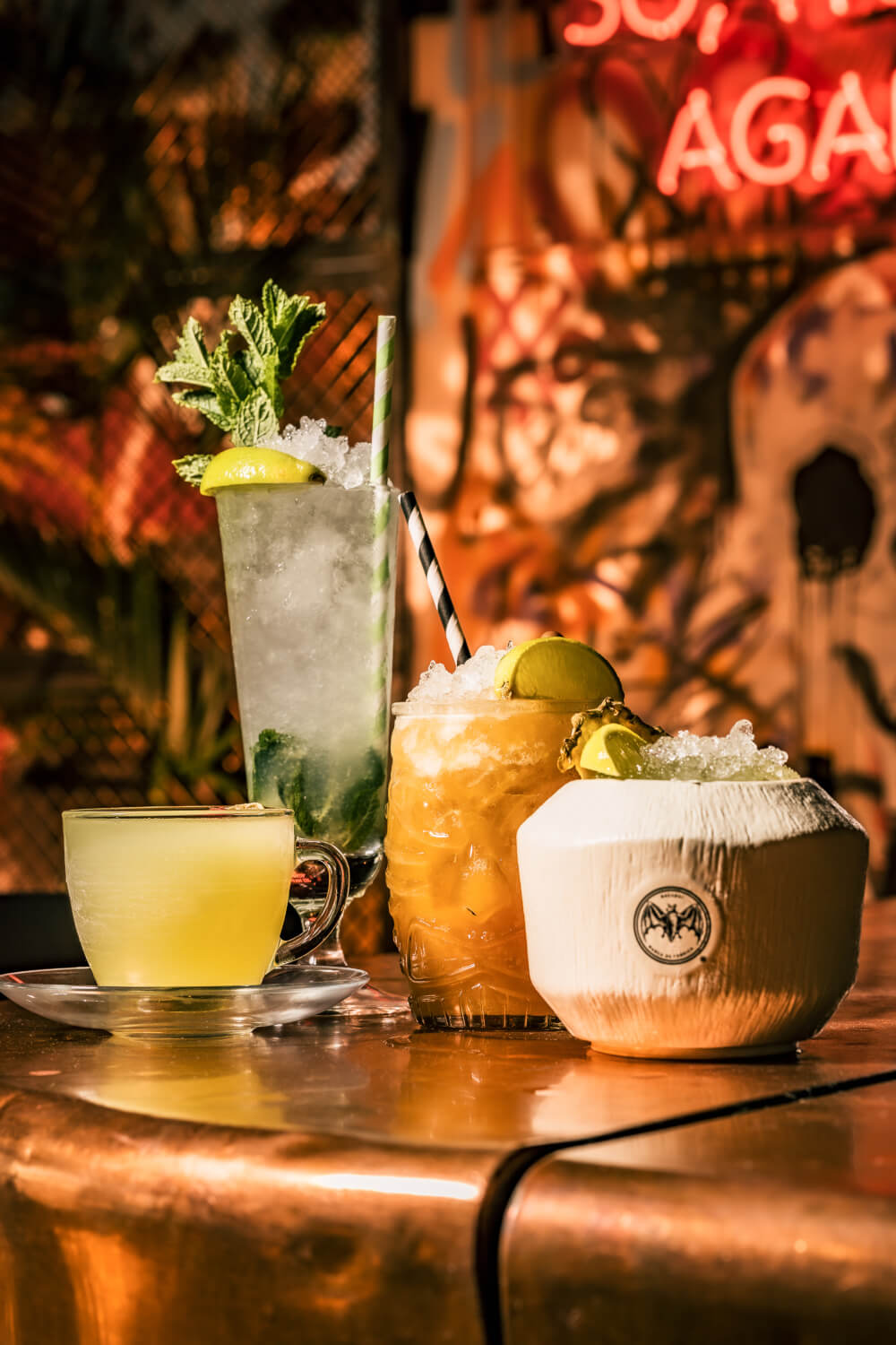 Four cocktails arranged on a bar: yellow, mojito, amber drink, white coconut cup with lime.