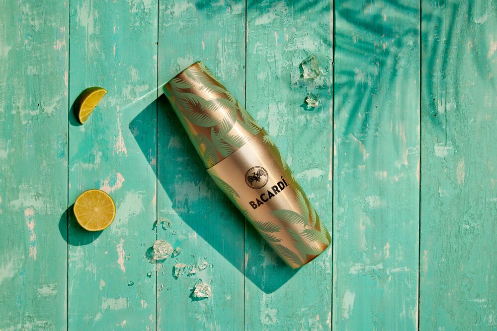 Gold BACARDÍ cocktail shaker with green palm leaf pattern, surrounded by lime slices and ice cubes.