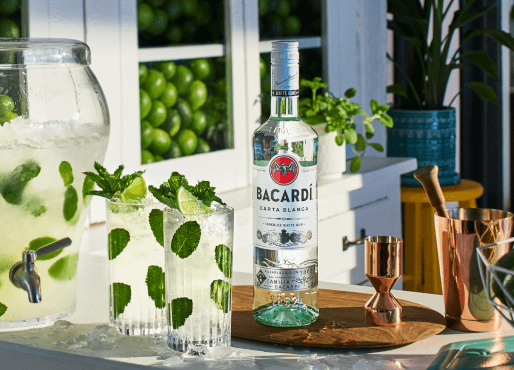 Bacardí Carta Blanca rum bottle with mojito ingredients, glasses, copper bar tools.