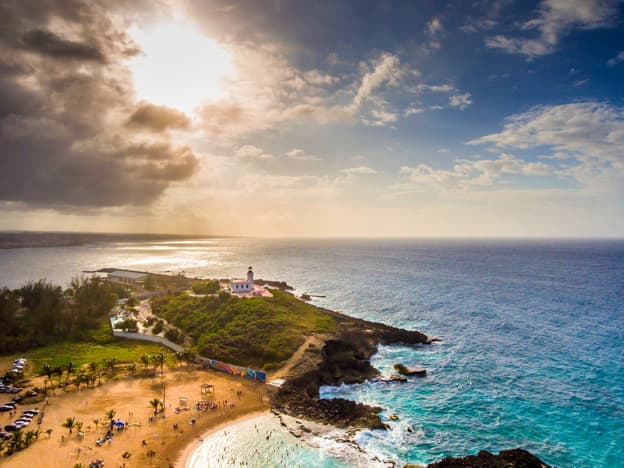 Aerial view of coastal landscape with sandy beach, lighthouse, palm trees, and blue ocean under partly cloudy sky.