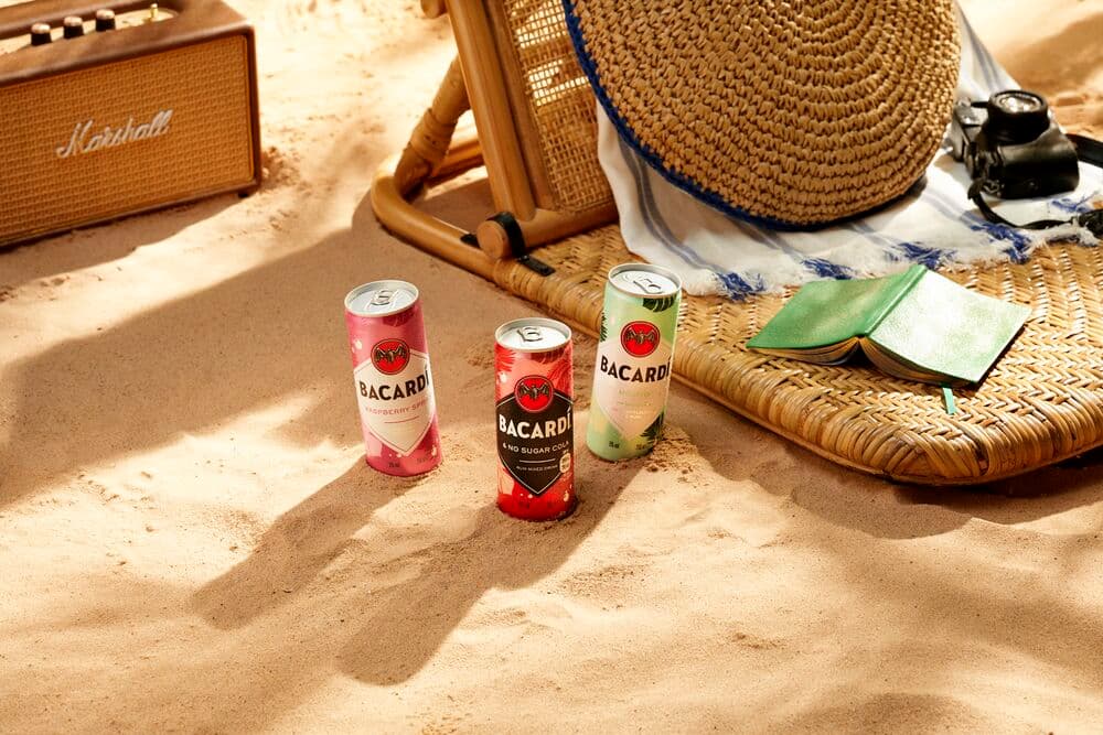 Three BACARDÍ cocktail cans positioned in sand with beach accessories like speaker, chair, hat.