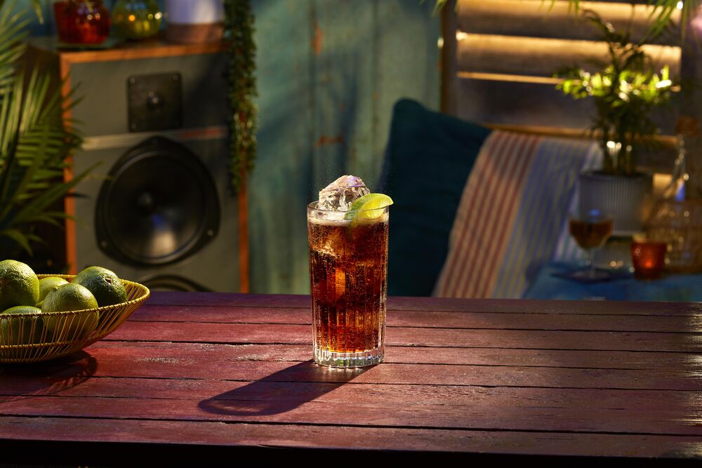 Dark cocktail with lime wedge in tall glass, accompanied by wire bowl of fresh limes.