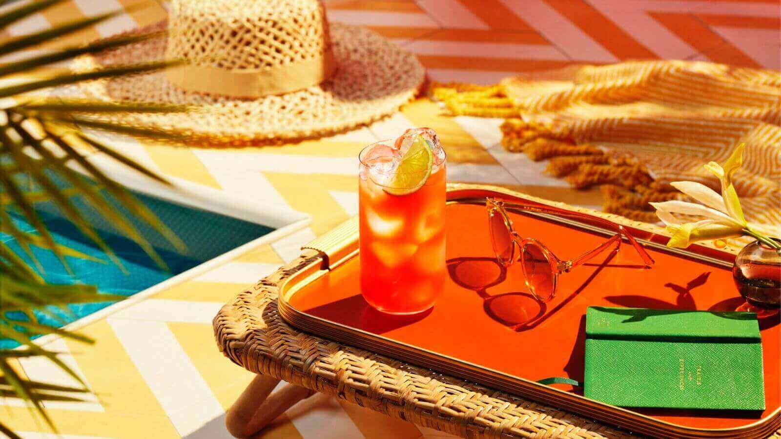 Red rum punch cocktail on orange tray with sunglasses, notebook near swimming pool.