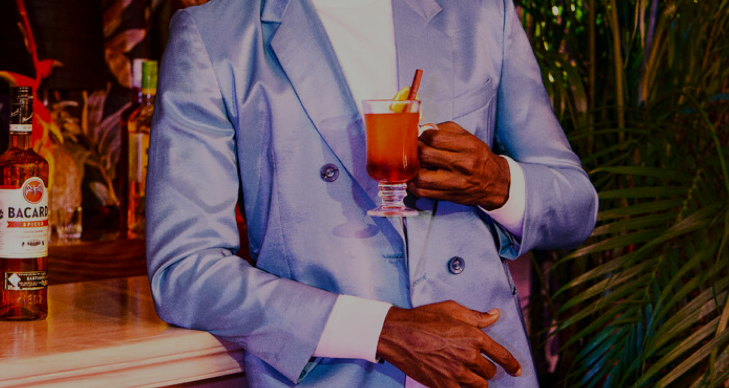 Person in light blue suit holding a warm-colored cocktail with cinnamon stick and citrus garnish.
