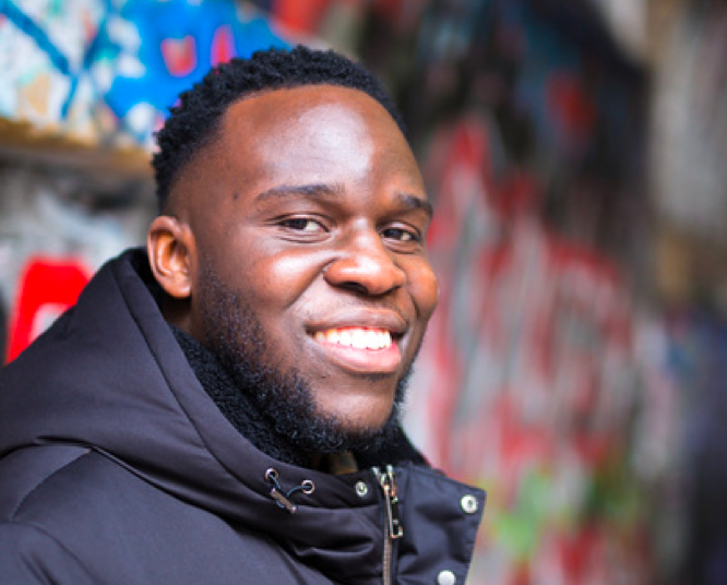 Person with a short beard smiling in a black puffer jacket against a blurred graffiti background.