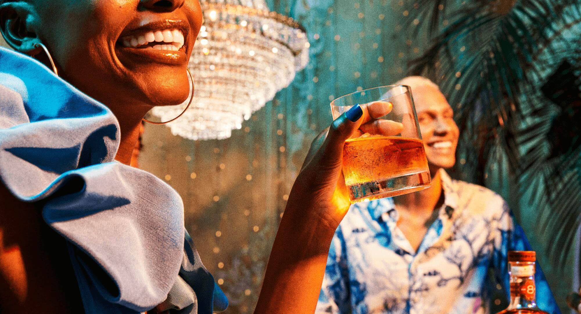 Person holding an amber cocktail glass at a festive gathering with palm leaves visible.