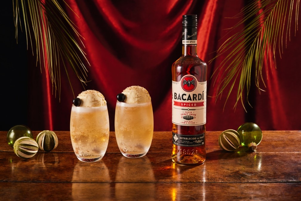 Bacardí Spiced rum bottle with two cream-topped cocktails, Christmas ornaments, dried citrus.